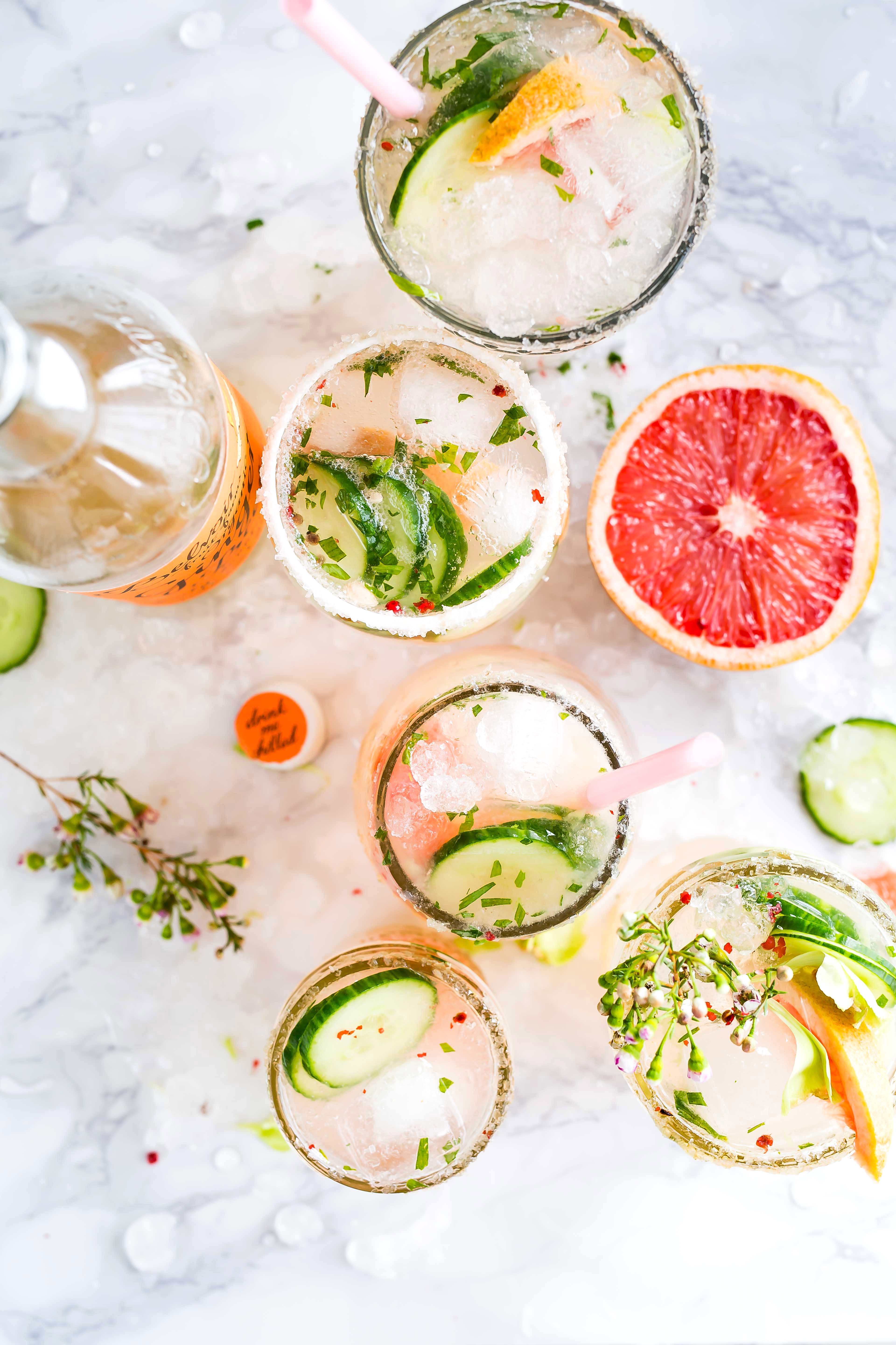 5 cocktails on a marble countertop with grapefruit, cucumbers, and flowers.