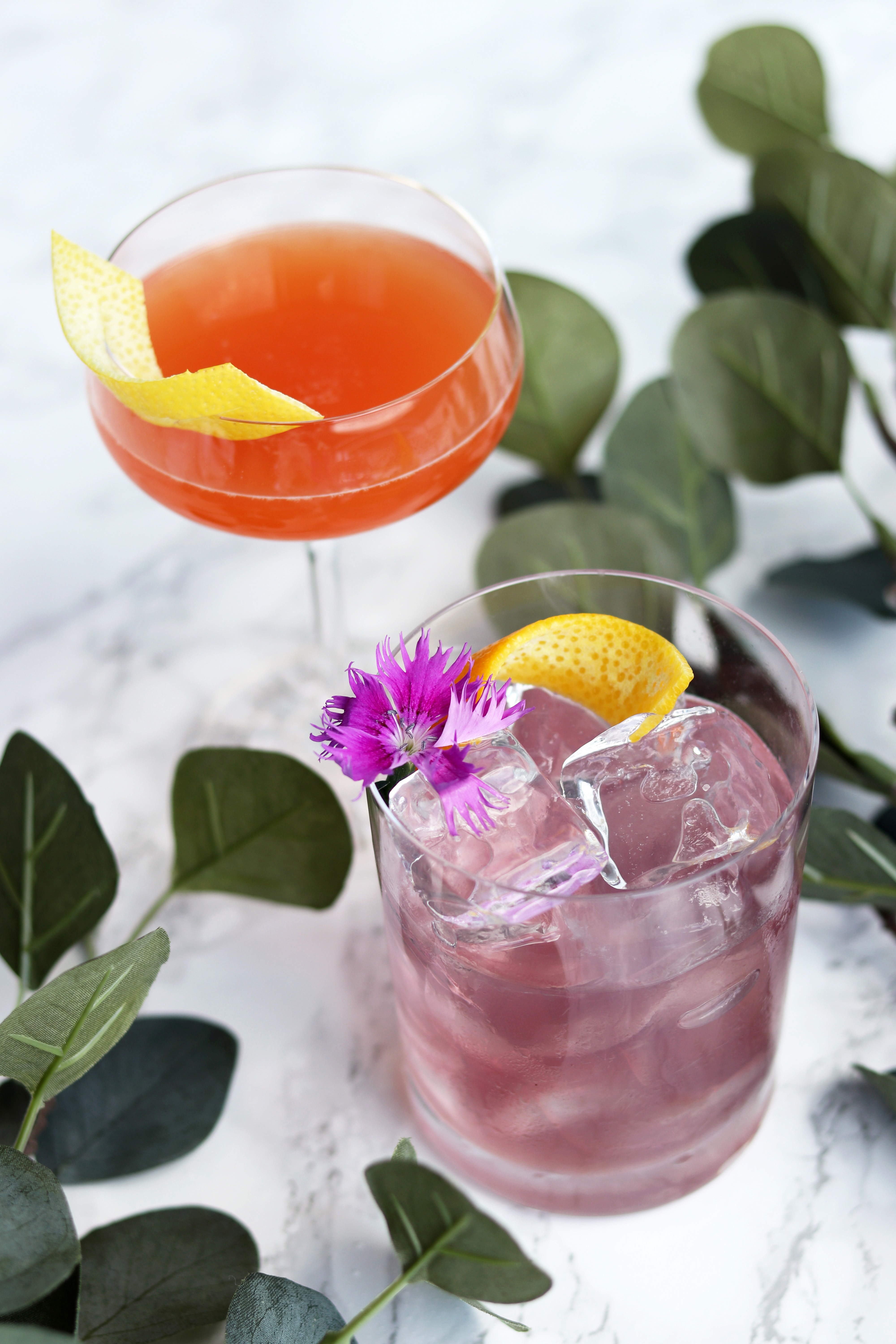 An orange cocktail with a lemon garnish and a purple cocktail with a lemon and flower garnish on a table.
